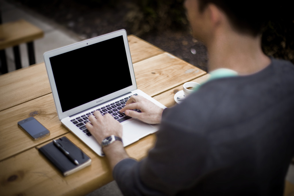 Man typing on a laptop on a wooden desk with a notebook and a coffee next to him.